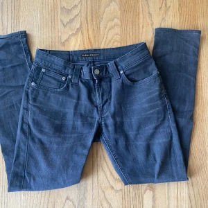 Nudie Jeans mörk grå - Säljer ett par grå jeans från Nudie Jeans Co med klassisk femficksdesign och snygga sömdetaljer på bakfickorna. Jeansen har normal passform och raka ben, perfekta till vardagslooken.