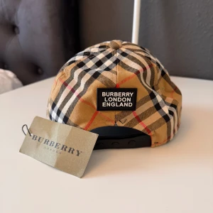 Rutig keps från Burberry - Säljer en klassisk rutig keps från Burberry med justerbar baksida. Kepsen har det ikoniska beige, svarta, vita och röda rutmönstret samt Burberry-logga bak. Perfekt accessoar för en trendig look.