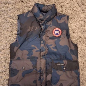Blå och grå camouflageväst från Canada Goose - Säljer en blå och grå camouflageväst från Canada Goose. Bara använd en gång efter att den inte passade. Västen är en present från början och har bara legat upphängd på en galge i garderoben. Västen har tryckknappar och ett tydligt Canada Goose-märke på bröstet. Perfekt för dig som vill sticka ut med en snygg och unik väst.