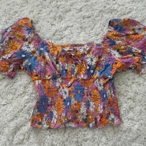 Blommig croppad blus med volang - Superfin blommig croppad blus i färgerna lila, orange, blå och vit. Toppen har korta puffärmar, volangdetalj vid halsringningen och smockad midja. Perfekt för sommarens alla tillfällen! 💞
