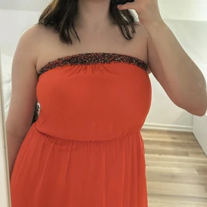 Röd axelbandslös långklänning med pärldetaljer - Säljer en röd axelbandslös långklänning med slits och vackra pärldetaljer längs överkanten. Klänningen har en luftig och flowy passform. Materialet känns lätt och svalt mot huden. Oanvänd. Prislappen finns kvar.