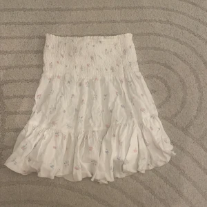 Vit blommig volangkjol - Supersöt vit kjol med små blommor i pastellfärger och volang nertill. Kjolen har smock i midjan för en bekväm passform och är kort i modellen. Perfekt för varma dagar 