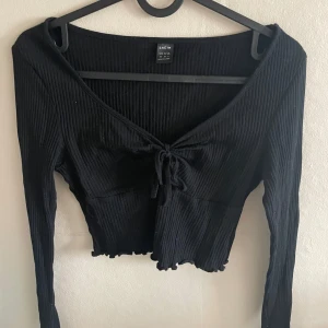 Svart ribbad croppad topp från Shein - Svart långärmad croppad topp från Shein med ribbad struktur och v-ringning. Toppen har knyt framtill och en lätt vågig nederkant. Perfekt för att skapa en trendig look.