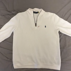 Vit långärmad tröja med dragkedja från Polo Ralph Lauren - Säljer en vit långärmad tröja från Polo Ralph Lauren med halv dragkedja och klassisk broderad logga på bröstet. Tröjan har en hög krage och är tillverkad i mjukt bomullsmaterial. Perfekt för en stilren och avslappnad look.