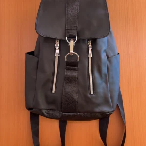 Svart ryggsäck med dragkedjor - Snygg svart ryggsäck med två silverfärgade dragkedjor framtill och justerbara axelband. Väskan har ett lock med spänne och snörning under locket för extra säkerhet. Perfekt för att bära med sig det viktigaste till skolan eller stan.