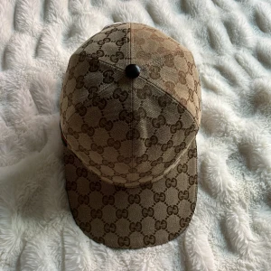 Beige keps från Gucci med GG-mönster - Säljer en beige keps från Gucci med klassiskt GG-mönster i brunt och en grön-röd rand på sidan. Kepsen har böjd skärm och justerbar rem baktill. Perfekt för dig som gillar lyxiga accessoarer med ikonisk design.
