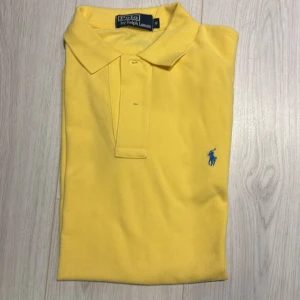 Gul pikétröja från Ralph Lauren - Snygg gul pikétröja från Ralph Lauren med klassisk krage och knappar framtill. Tröjan har det ikoniska blå broderade logotypen på bröstet. Perfekt för en stilren och sportig look. Aldrig använd bara legat i garderoben! Köptes för 1399 i Ralph Lauren egna butik utomlands för längesedan. Skriv i DM vid frågor!