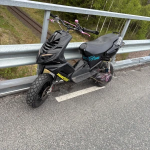 Spääd - Säljer en svart moped med rosa handtag och bromsdetaljer. Sadel med Coca-Cola-tryck och sidodekal med OnlyFans-logga. Mopeden har grova däck och sportigt utseende. Vissa delar och motordelar syns lösa på marken. Köp den för 9000 finns i värnamo de e bara o skriva fps med lite trim delar 
