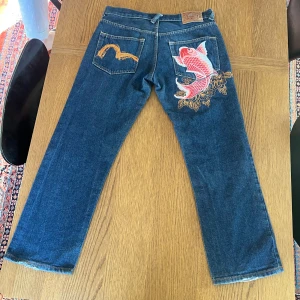 Evisu jeans med broderad koi-fisk - Unika blå jeans från Evisu med en stor broderad röd koi-fisk och gulddetaljer på bakfickan. Klassisk femficksmodell med raka ben och knappar i gylfen. Snygg kontrastsöm och ikonisk Evisu-logga på bakfickan. Lägg märke till att sömmarna längst när på byxan har börjat fransa.