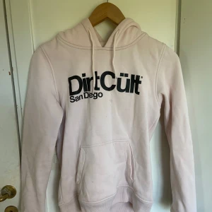 Ljusrosa hoodie från Dirt Cült - Säljer en ljusrosa hoodie från Dirt Cült San Diego med svart tryck framtill. Tröjan har huva med dragsko och en stor magficka. Tillverkad i 65% bomull och 35% polyester. Perfekt för en avslappnad stil.