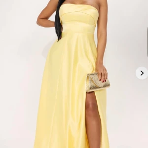Gul axelbandslös långklänning med slits - Säljer en elegant gul axelbandslös långklänning med vacker drapering och hög slits framtill. Klänningen har en figurnära överdel och markerad midja, samt dekorativa volangdetaljer baktill. Perfekt för speciella tillfällen!