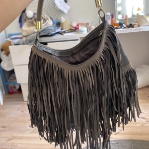 Grå axelväska med fransar - Säljer en snygg grå axelväska med långa fransar och guldfärgade metalldetaljer. Väskan har dragkedja upptill och är rymlig med innerfack. Perfekt för dig som vill ha en trendig accessoar med boho-vibe.