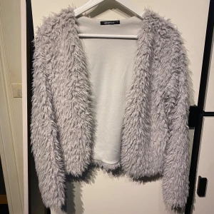Ljusgrå pälsjacka från Gina Tricot - Säljer en fluffig ljusgrå pälsjacka från Gina Tricot. Jackan har lång ärm och öppen front utan synliga knappar eller dragkedja. Perfekt för dig som vill sticka ut med en mysig och trendig look.