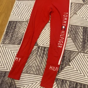 Röda leggings från Tommy Hilfiger - Snygga röda leggings från Tommy Hilfiger med vit sidoremsa och stort logotryck längs benet. Bekväm passform och elastiskt material, perfekta för en sportig och trendig look.