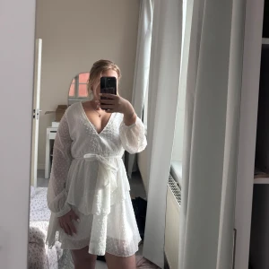 Vit omlottklänning med volang och knytband - Superfin vit omlottklänning med volangdetaljer och knytband i midjan. Klänningen har långärmade, lätt transparenta ärmar och ett diskret prickigt mönster. Perfekt för sommarens alla tillfällen! Har tecken på användning kom privat för fler bilder. 