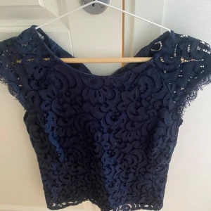 Mörkblå spetsblus från Holly & Whyte - Säljer en mörkblå blus från Holly & Whyte med vackert spetsmönster. Blusen har korta ärmar och rund halsringning. Perfekt för dig som vill ha en elegant och stilren topp med fina detaljer.