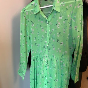 Grön blommig långklänning med krage - Säljer en söt grön långklänning med små vita och rosa blommor. Klänningen har lång ärm, klassisk skjortkrage och knappar framtill. Perfekt för vår och sommar!