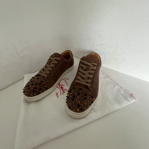 Unika brun beiga Christian Louboutin skor - Säljer ett par bruna sneakers från Christian Louboutin med ikoniska röda sulor. Skorna har snörning och är dekorerade med metallnitar på tå och häl. Ovandelen är i mocka och sulan är vit med röd undersida. Det finns ingen skada på den röda sular men det är smuts runt på det vita alla bitar sitter på och det finns 3 extra nitar. Tveka inte på att höra av dig vid frågor!