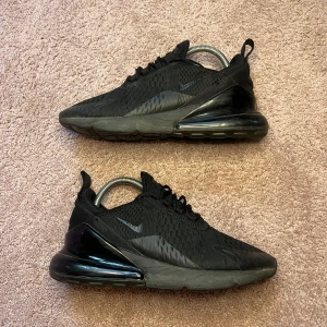 Svarta Nike Air Max 270 sneakers - Säljer ett par helsvarta Nike Air Max 270 sneakers med mesh-överdel och synlig Air Max-dämpning i hälen. Skorna har snörning och en sportig, modern design med diskret Nike-logga på sidan och tungan.