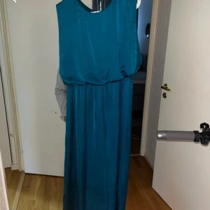 Blå bal klänning i satin - Säljer en blå långklänning i satin med ärmlös design och rund halsringning. Klänningen har en öppen detalj i ryggen och ett elegant, flytande fall. Perfekt för dig som vill ha en stilren och enkel look.