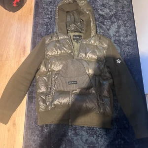 Olivgrön pufferjacka med huva från Bvaldi - Säljer en olivgrön pufferjacka från Bvaldi med huva och dragkedja. Jackan har quiltad framsida i glansigt material och stickade ärmar. Märkespatch på ena ärmen och två fickor med dragkedja framtill.