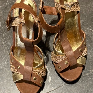 Bruna sandaler med klack från H&M - Säljer ett par bruna sandaler med hög klack från H&M. Skorna har remmar i brunt och guldigt syntetmaterial, dekorerade med nitar och spänne vid ankeln. Snyggt mönstrade detaljer och öppen tå ger en trendig look. Upplever storleken som 38. 
