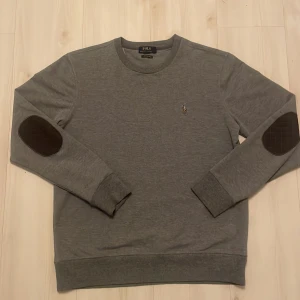 Grå sweatshirt från Polo Ralph Lauren - Säljer en stilren grå sweatshirt från Polo Ralph Lauren med bruna armbågslappar. Tröjan har långa ärmar och en liten broderad logotyp på bröstet. Perfekt för en avslappnad stil.