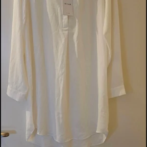 Vit långärmad tunika från Vila - Säljer en stilren vit tunn tunika från Vila i nyskick, lappen kvar. Tunikan har en rak passform och är tillverkad i ett lätt material som ger en luftig känsla. Perfekt att styla till olika outfits.