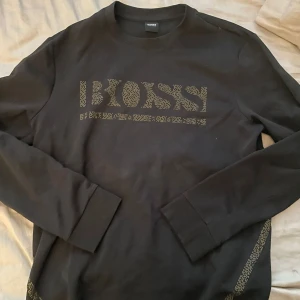 Svart långärmad tröja från BOSS - Säljer en svart långärmad tröja från BOSS med logga i mönstrad stil framtill. Tröjan har rund halsringning och är tillverkad i mjukt material. Perfekt för en stilren och avslappnad look.