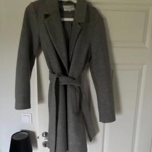 Grå kappa - Säljer en stilren grå kappa från Vero Moda. Kappan har lång ärm, klassisk krage och knyts med bälte i midjan för en snygg siluett. Perfekt för dig som vill ha en tidlös och elegant look. Skriv för fler bilder 🩷