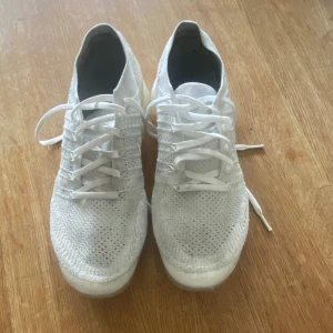Nike Air Vapormax Flyknit vita sneakers - Säljer ett par vita Nike Air Vapormax Flyknit sneakers med stickad ovandel och genomskinlig sula. Skorna har snörning och är tillverkade i lätt och ventilerande material. Perfekta för dig som gillar stilrena och sportiga skor.