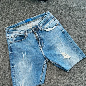 Blå jeansshorts från Zara Man - Säljer ett par blå jeansshorts från Zara Man med slitna detaljer och råa benslut. Klassisk femficksmodell med dragkedja och knapp. Perfekta för en avslappnad stil under varma dagar.