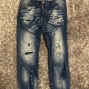 Blå jeans från Dsquared2 - Säljer ett par blå jeans från Dsquared2 med slitna detaljer och färgstänk. Byter gärna mot andra jeans i lite mindre storlek De har en straight passform och klassisk femficksdesign. Perfekta för en edgy look! Använda Max 2-3 gånger 