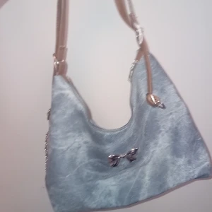 Grå axelväska med rosettdetalj - Säljer en snygg grå axelväska med beige axelrem och silverfärgad kedjedetalj. Väskan har en liten rosett framtill och är rymlig med ett stort huvudfack. Perfekt för dig som vill ha en stilren och trendig accessoar.