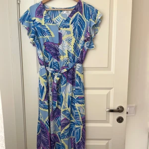 Blå och lila mönstrad långklänning med volangärm - Färgglad långklänning med tropiskt bladmotiv i blått, lila och gult. Klänningen har volangärmar, knappar framtill och ett matchande knytskärp i midjan. Perfekt för sommarens alla tillfällen.