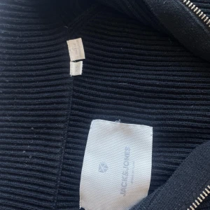 Svart ribbad tröja med dragkedja från Jack & Jones - Svart långärmad ribbad tröja från Jack & Jones med halv dragkedja framtill. Hög krage som kan vikas ner och stilren design som passar till många tillfällen. Perfekt för dig som gillar enkel och snygg stil.