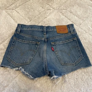 Levi's jeansshorts med fransar - Säljer ett par klassiska blå jeansshorts från Levi's med fransar nertill och slitna detaljer framtill. Shortsen har låg midja, fem fickor och knäppning med knappar. Perfekta för sommaren!