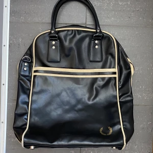 Svart Handväska från Fred Perry - Säljer en svart Handväska från Fred Perry med krämfärgade detaljer och dragkedja. Väskan har dubbla handtag, en yttre ficka och klassisk Fred Perry-logga i guld. Perfekt för att bära med sig det viktigaste till skolan eller stan. Snabb Leverans!