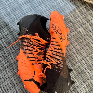 Puma Future orange fotbollsskor - Säljer ett par grymma Puma Future fotbollsskor i orange och svart. Skorna har snörning, högre strumpliknande skaft och coolt mönster med svarta detaljer. Ovandelen är i syntetmaterial och sulan har fasta dobbar för bästa grepp på planen.