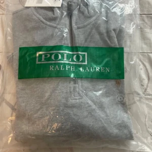 Grå half zip tröja från Polo Ralph Lauren - Snygg grå långärmad tröja från Polo Ralph Lauren med half zip och klassisk broderad logga på bröstet. Tillverkad i mjuk bomull, perfekt för en clean och stilren look. Ribbstickade muddar och hög krage ger extra edge.