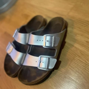 Birkenstock sandaler silver strl 40 - Snygga Birkenstock sandaler i metallic silver med två breda remmar och justerbara spännen. Ovandel i syntet, fotbädd i mocka och kork, samt svart mönstrad gummisula. Perfekta för sommaren och riktigt bekväma att gå i.