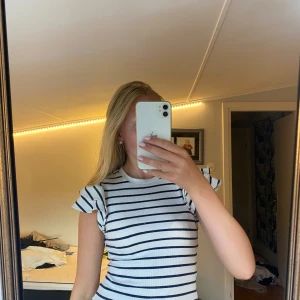 Randig topp med volangärmar Holly & White - Superfin ribbad topp från Holly & White i vitt med svarta ränder. Den har korta volangärmar och rund halsringning. Perfekt för dig som gillar en söt och stilren look med lite extra detalj på ärmarna.