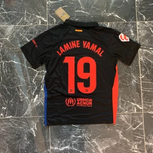 FC Barcelona Lamine Yamal tröja Nike - Säljer en svart FC Barcelona fotbollströja med Lamine Yamal och nummer 19 på ryggen. Tröjan har röda och blå detaljer på sidorna, klubbmärke på bröstet och UNHCR-logga bak. Tillverkad i lätt och ventilerande material, perfekt för match eller träning.