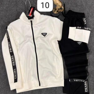 Vit och svart tracksuit från Prada - Snygg tracksuit från Prada i vitt och svart med svarta detaljer och logga på både jacka och byxa. Jackan har dragkedja och svarta ränder med Prada-tryck längs ärmarna. Byxorna har resår i midjan och matchande logga och ränder.