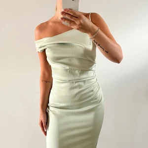 Ljusgrön långklänning med slits - Snygg ljusgrön långklänning i satinliknande material med offshoulder och smala axelband. Klänningen har figurnära passform, draperad detalj vid midjan o slits. Klänningen kommer ifrån Adoore och använts vid en tillfälle 