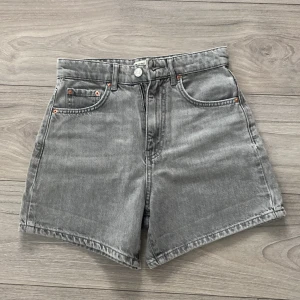 Gråa jeansshorts från Gina Tricot - Jättefina gråa jeansshorts från Gina Tricot med hög midja och klassisk femficksdesign. Shortsen har raka ben, bälteshällor och dragkedja med knapp framtill. Perfekta till sommaren för en chill och trendig look.