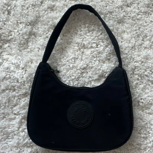 Svart axelväska XO Stay Kind - Snygg svart axelväska med rundad form och bred axelrem. Väskan har en dragkedja upptill och ett stilrent gummimärke framtill med texten 'XO Stay Kind'. Perfekt storlek för det viktigaste och enkel att matcha till olika outfits.