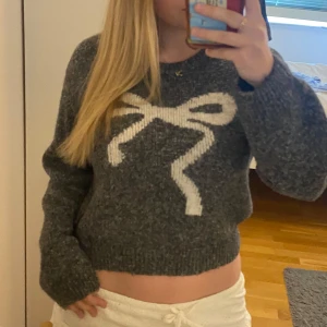 Mörkgrå stickad tröja med rosett - Mysig mörkgrå stickad tröja med en stor vit rosett framtill. Knappt använd. 