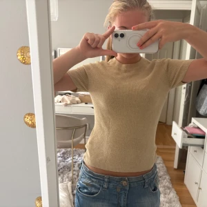 Guldig topp - En guldig stickad topp från Gina Tricot med korta ärmar och rund hals. Toppen har en ribbad struktur och sitter snyggt på kroppen. Perfekt att styla med jeans eller kjol för en clean look.