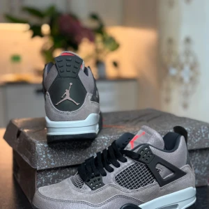 Nike Air Jordan 4 Taupe Haze sneakers - Nike Air Jordan 4 Taupe Haze med grå mocka, svarta detaljer och meshpaneler. Snygg kontrast med röd insida och Jumpman-logga på häl och sula. Klassisk siluett med svart snörning och vit/svart sula. Perfekta för dig som vill sticka ut med stilrena sneakers.